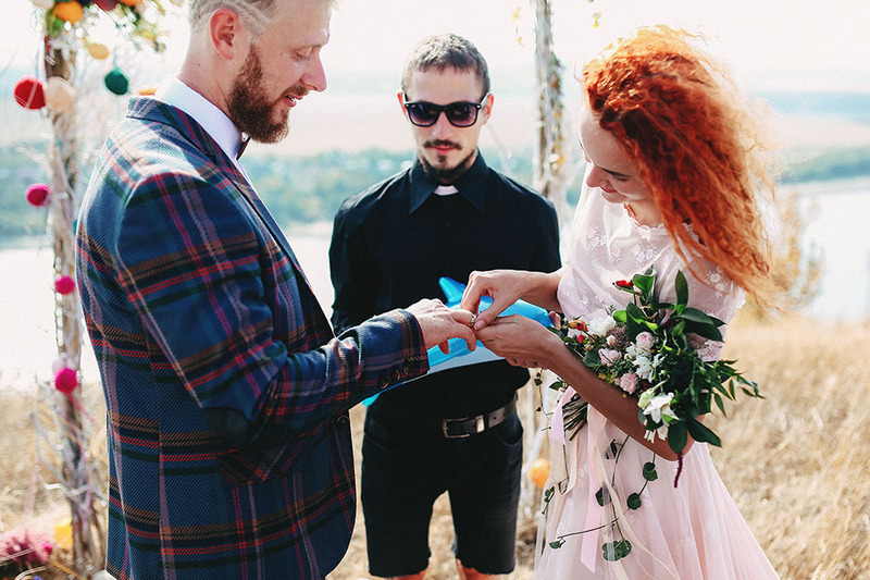 A bride places the ring on the grooms finger while the wedding officiant oversees the exchange, outdoors in front of a colorful wedding arch