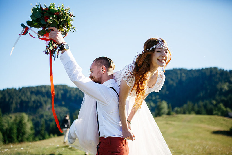A groom holds up the wedding bouquet as he carries a bride over his shoulder laughing on the wedding day, they are outdoors in a field on a sunny day, with a bright blue sky 