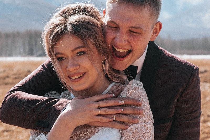 Newlyweds laugh at their outdoor wedding ceremony, the groom has his eyes closed and his mouth open in a big laugh, embracing the bride from behind. She looks amused. 