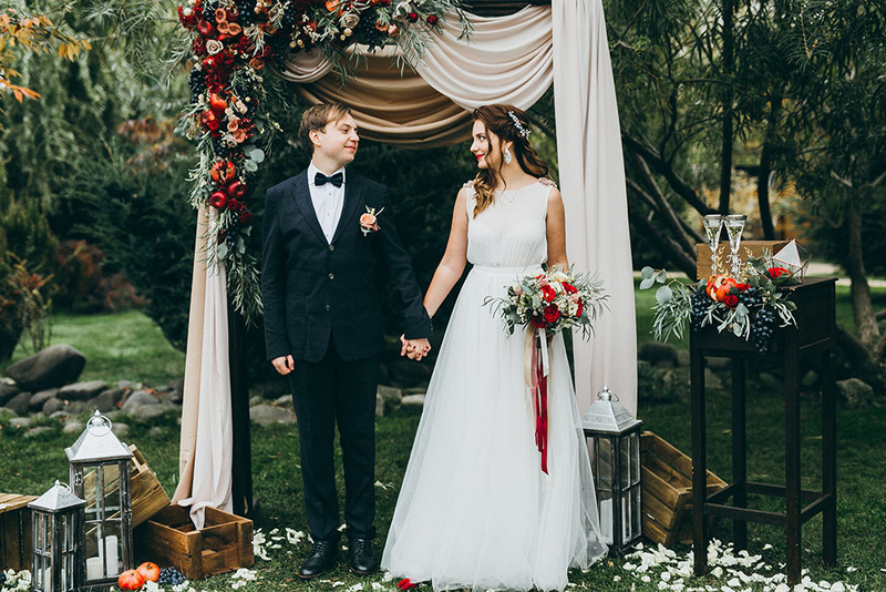 A happy couple of newlyweds hold hands outdoors next to the wedding arch, decorated with seasonal flowers, with a forest behind them