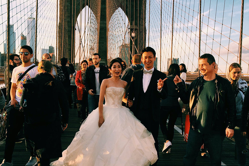 Photo of a couple getting married on the Brooklyn Bridge, with NYC in the background, surrounded by friends