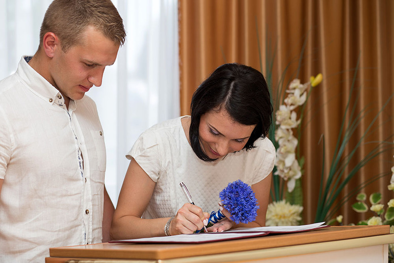 A young couple of newlyweds take turns signing their marriage license following the ceremony, the bride holds a bouquet while she signs