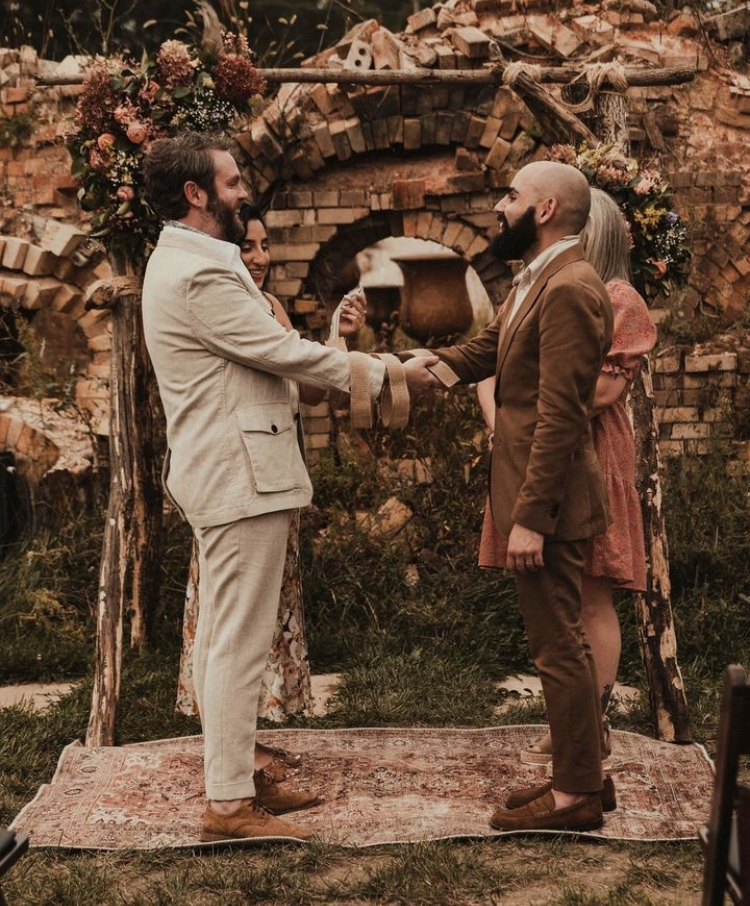 Photo via Liz Rae Weddings, two men stand facing each other, holding hands with a handfasting ribbon wrapped around their wrists during a wedding ceremony 