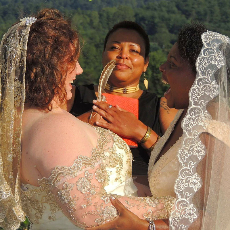 Two brides link arms and smile happily during their wedding ceremony, while their wedding officiant smiles and holds up a feather 