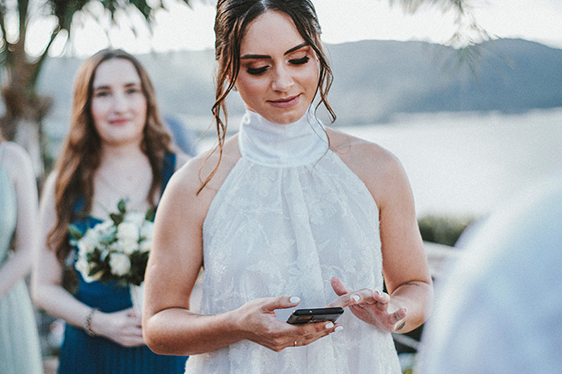 Bride looks at her cellphone, scrolling, during the wedding ceremony. Behind her is a bridesmaid, holding flowers