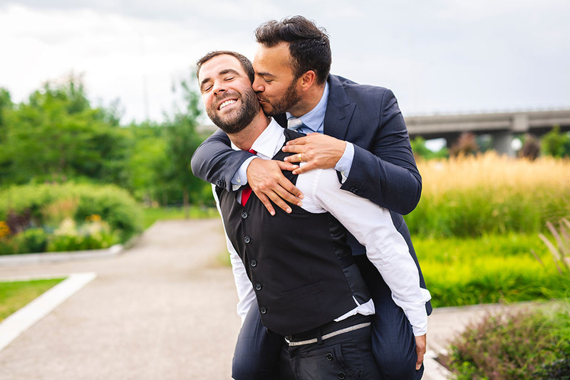 Two grooms embrace, smile, kiss on cheek outdoors on their wedding day