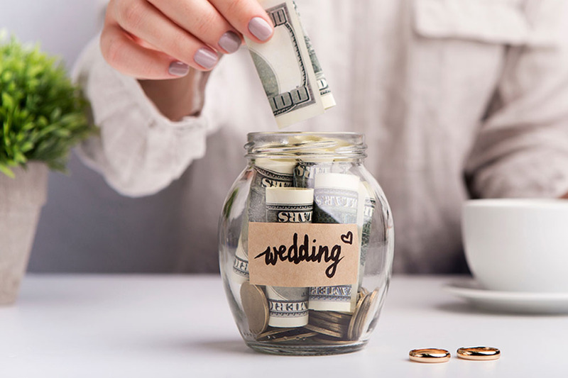 Close up photo of a woman with painted nails sticking a folded up 100 dollar bill into a jar with the label "Wedding"