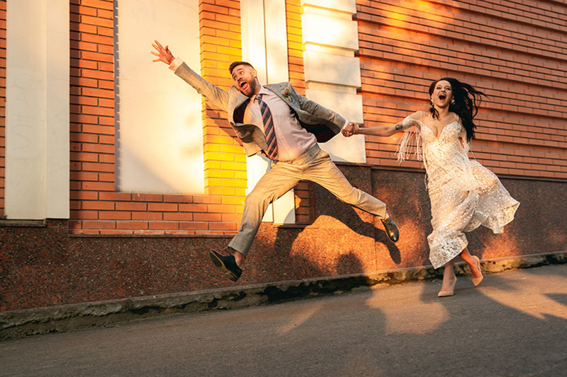 Newlyweds pose for a photo while jumping in the air outside a city building, wearing their wedding outfits and making happy faces. 