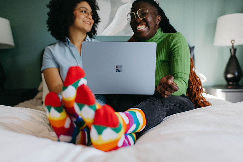 A happy couple lay in bed with a laptop, writing wedding vows using ChapGPT. They are wearing casual clothes and brightly colored socks, and smiling and laughing together. 