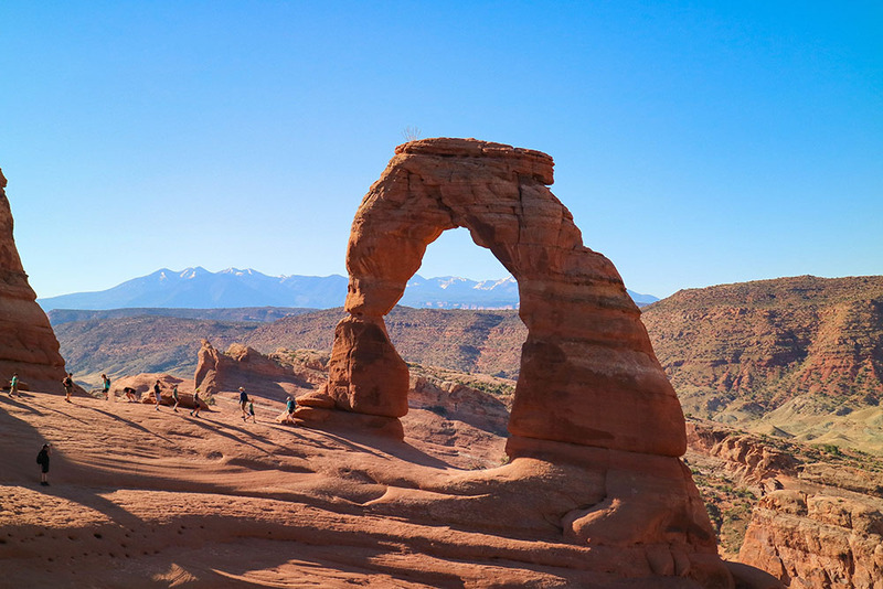 Photo shows a red rock arch against a blue sky, with people walking below on the rocks. They seem tiny in comparison to the large rock formation. Taken at Arches National Park