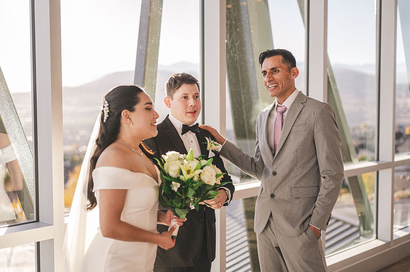 Photo shows a male wedding officiant in a suit standing next to a bride and groom on the wedding day, wearing a white wedding dress and a suit. 