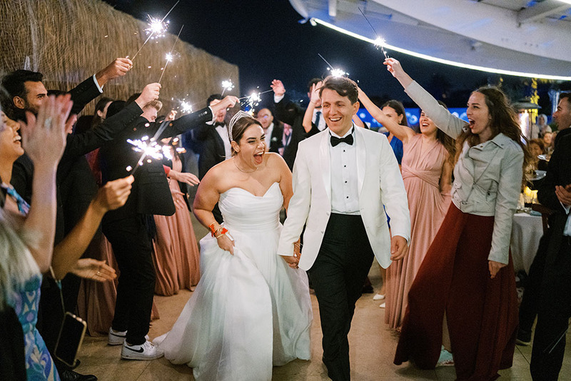 Newlyweds walk under an 'arch' made by friends and family holding sparklers. It's night, and the sparklers light up the couple's faces, as they laugh and smile happily