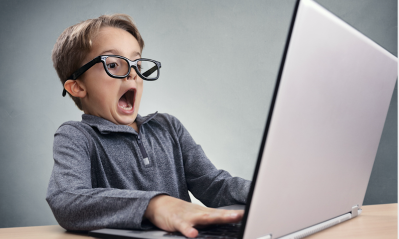 Funny image of a young boy in oversized glasses making a face of surprise at a computer screen