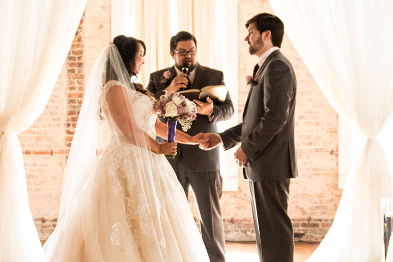 Photo is in slightly overexposed cream and rust tones, soft sepia tones, and shows a bride and groom holding hands in front of a wedding officiant, who holds a microphone and looks down at the wedding script as he performs the ceremony