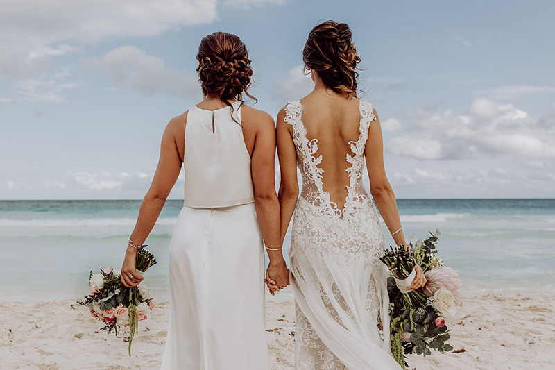 Two brides hold hands on the beach in their wedding dresses, their backs are toward the camera and they are looking out at the beautiful ocean view