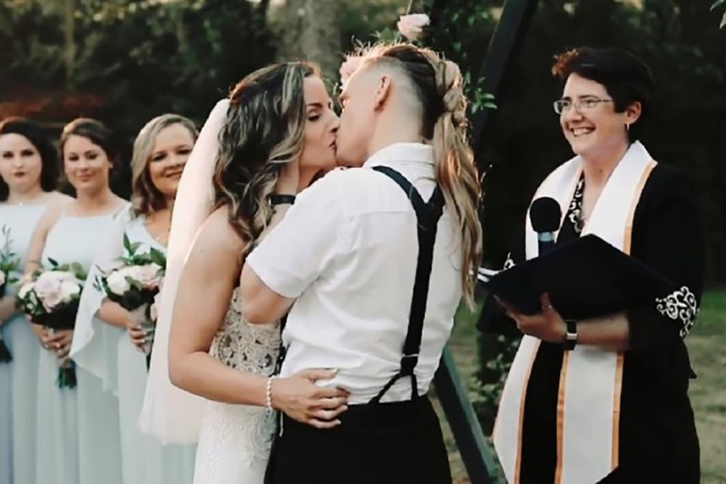 Two brides kiss on their wedding day while friends and the minister watch, smiling, after they're declared married