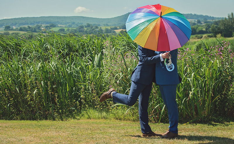 Grooms kiss outdoors behind a rainbow colored umbrella on their wedding day, in the background are rolling hills and farm land