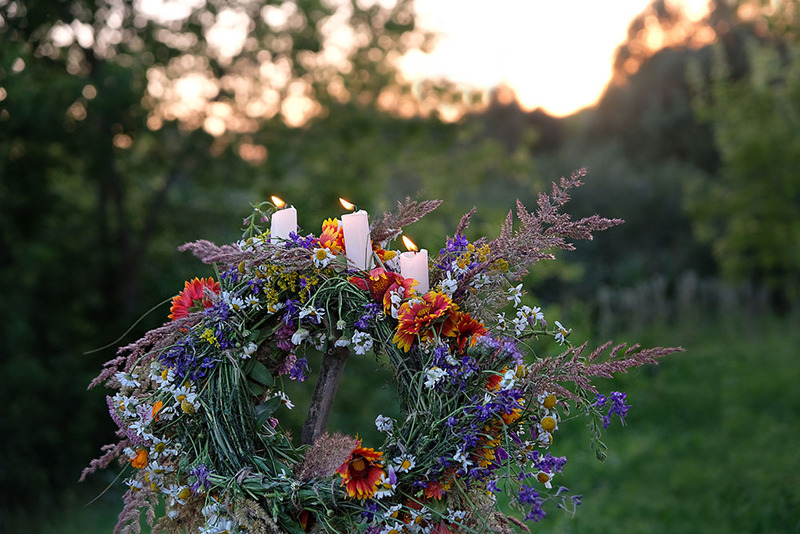 Flower wreath with three ceremonial white candles sits outdoors surrounded by lush green trees at sunset