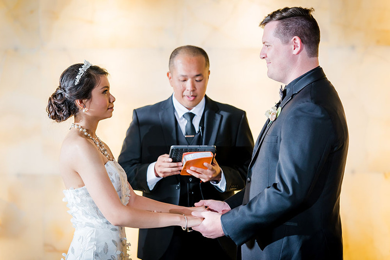 A wedding officiant performs a ceremony for a bride and groom, reading from the ceremony script while the couple hold hands