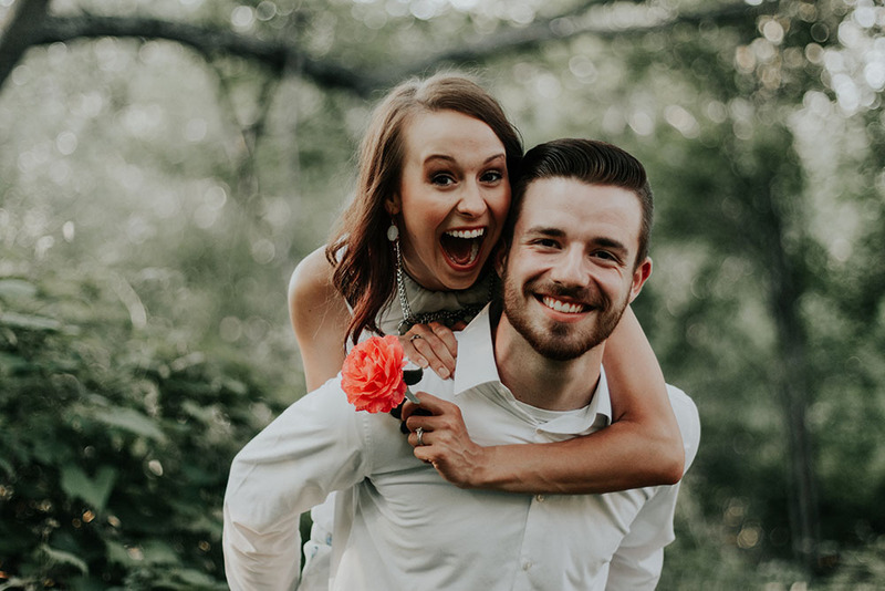 Excited newlyweds, the bride has her arms wrapped around the groom from behind as they smile at the camera