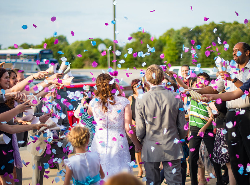 Taken from behind the newlywed couple as they walk towards friends and family tossing blue and purple petals as confetti