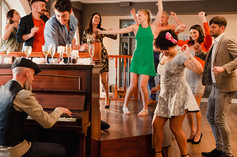 Friends dance at the reception to a song played by an older man on piano, wearing brightly colored casual party clothes following a solo wedding ceremony