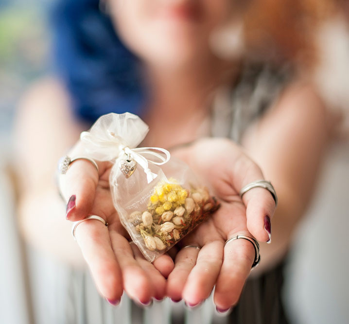 A solo bride holds out a pouch of herbs, close up photo of these herbs and her hands, she's wearing multiple silver rings and her blue hair and smile are out of focus in the background