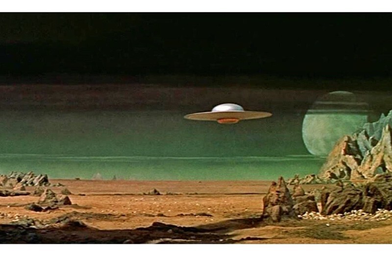 Screenshot from the vintage sci fi film Forbidden Planet shows the surface of an otherworldly planet with a distant planet in the sky with a celestial ring