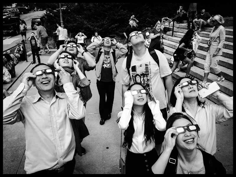 a group of people looking up at an eclipse, smiling, with eclipse glasses on