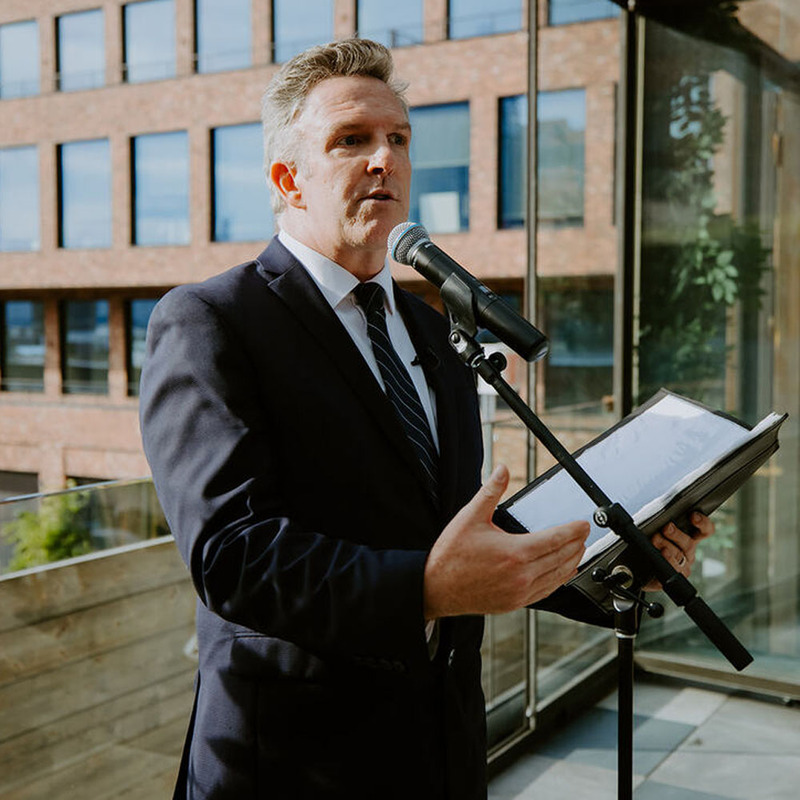 Christopher Shelley performs an outdoor wedding, he's speaking at a microphone holding the wedding ceremony script