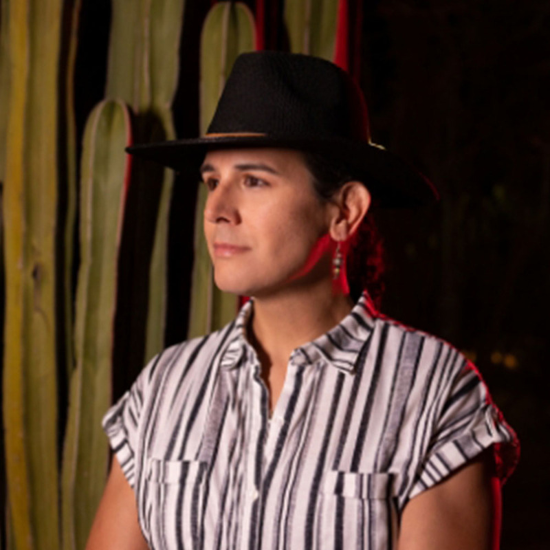 Don Mamone poses in front of cactus wearing a black hat and striped shirt, looking off to the side