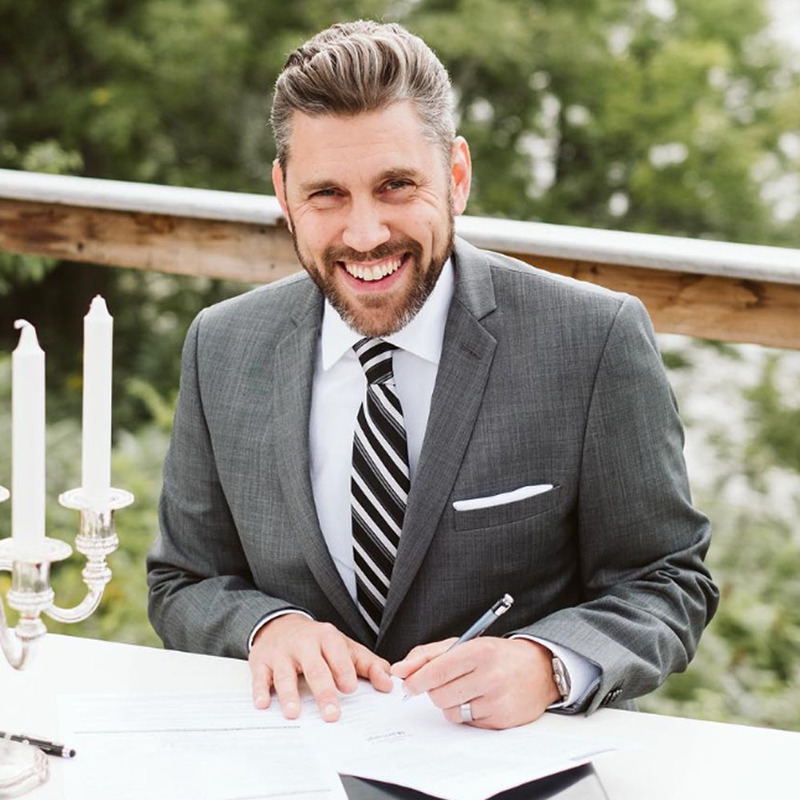 Mark Allan Groleau smiles at the camera wearing a sharp suit and tie while signing marriage documents following an outdoor wedding ceremony