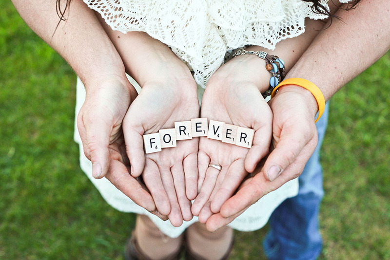 Close up of a man and woman holding their hands out, on top of each other, holding scrabble pieces that spell 'forever'