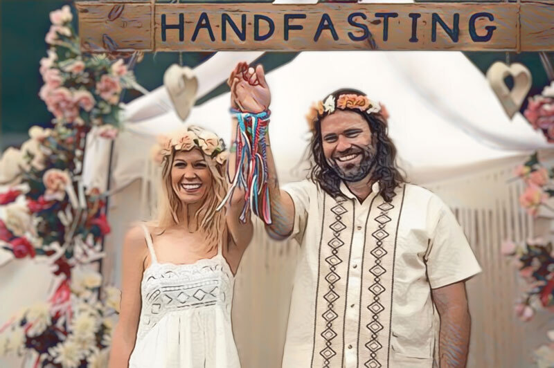 stylized photo of a couple with their hands raised, holding hands, tied at the wrist with ribbons. They are dressed in casual hippy style clothing in front of a wedding arch covered in pink roses, with the word 'handfasting' carved into the top of the arch