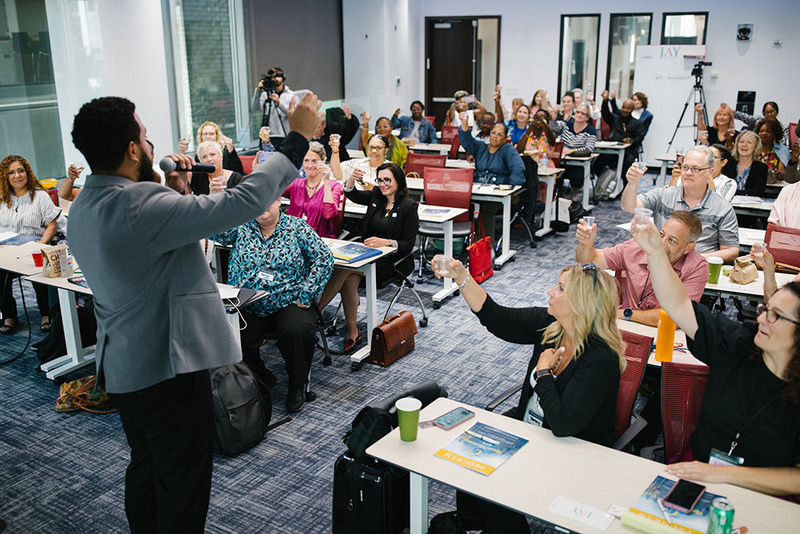 Officiant Jimmie Berguin leading attendees in a toast of chocolate candies during an Officiant Accelerator wedding officiant training event in New York City, Jimmie stands at the front of the room, the photo is taken from behind him, looking out into the room filled with smiling guests sitting down at long tables and holding up cups with candy in them