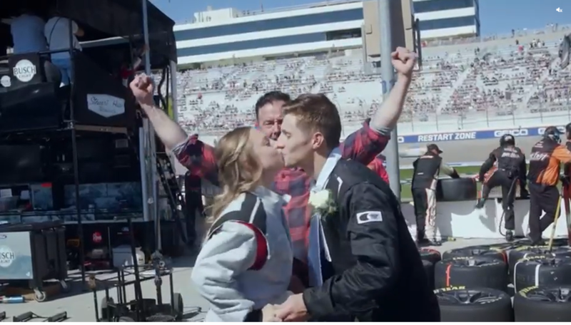 Newlyweds Tori and Nick kiss following their fast wedding ceremony during a NASCAR race in Las Vegas in October, the Officiant raises his arms excitedly, behind them is a stadium filled with fans