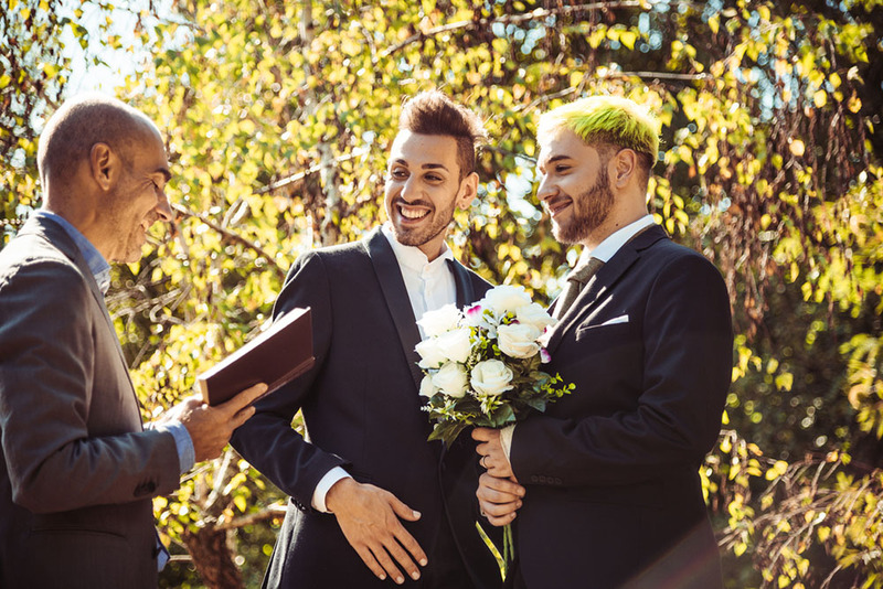 A wedding officiant performs a beautiful outdoor ceremony for two grooms, they are all wearing suits and smiling, its a sunny day