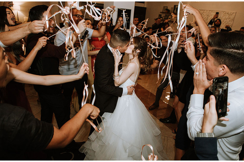 Newlyweds kiss while friends and family gather around them smiling and cheering, with ribbons