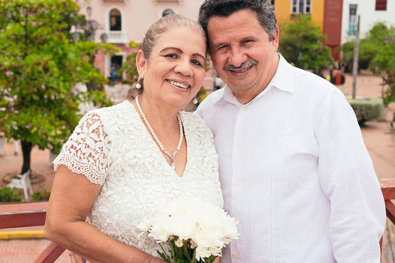 Older adult newlyweds pose for a photo outdoors on their wedding day, leaning together smiling, the bride holds a small bouquet