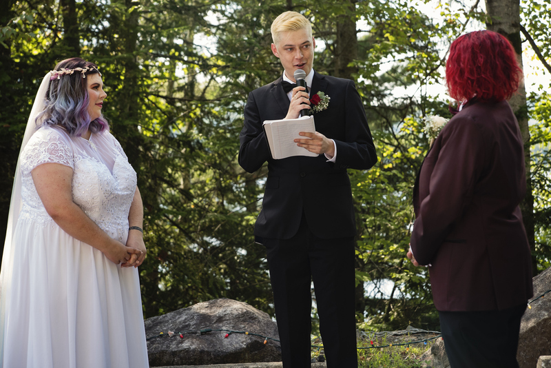 A sober wedding officiant performs an outdoor wedding in front of a beautiful green forest, the two brides stand on either side of him, smiling as he reads the ceremony script