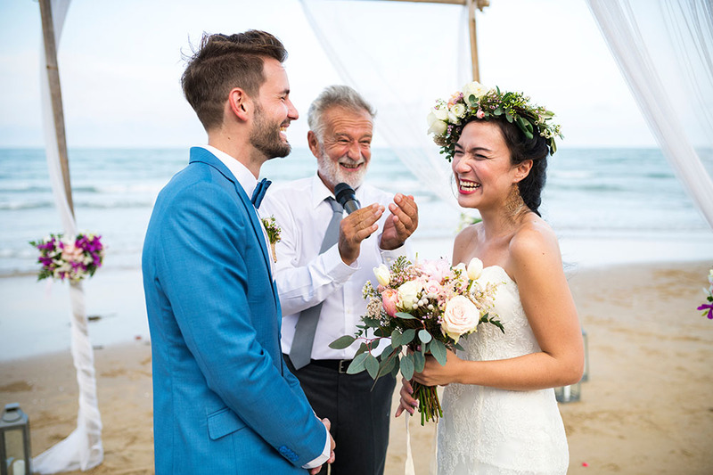 An older man officiates a wedding on a beach, standing with the bride and groom. Everyone is laughing and wearing formal wedding attire, in the background is the ocean