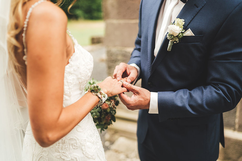 Close up photo shows a groom placing a wedding ring on the bride's ring finger during a wedding ceremony ring exchange. The bride wears a white beaded wedding gown with thin straps, and has long blonde hair. The groom wears a dark blue suit jacket, white button up shirt, and has a white rose in his lapel.