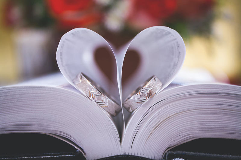 A book lays open on a table. Two pages are folded in towards the spine, creating the shape of a heart. Inside each curve of the 'heart' is a wedding ring.