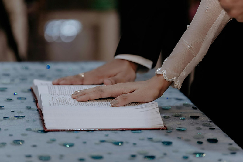 CLose up photo shows a bride and groom resting their hands, palm down, on an open Holy Bible during a religious CHristian wedding ceremony