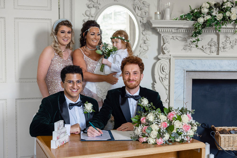 Two grooms sit at a small table signing their marriage license during the wedding unity ceremony. Behind them, two attendants in pink dresses watch and smile, one of them is holding a young flower girl