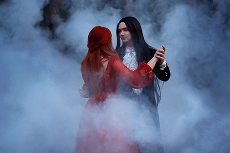 A young gothic alternative couple dance outdoors in a forest at night, surrounded by mystical fog. The bride wears a flowing red dress, and the groom has long dark hair and a dark wedding suit.