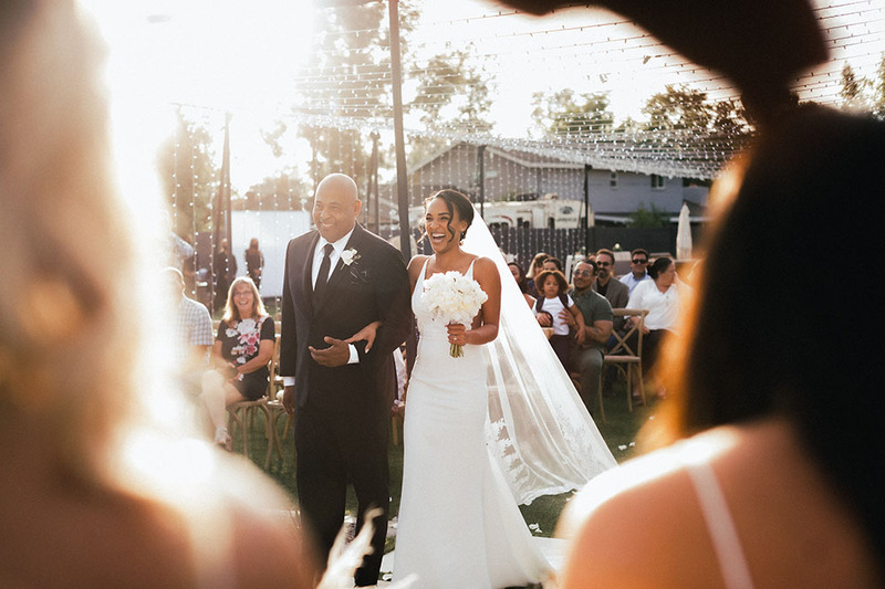 Outdoor wedding ceremony, bride walks down the aisle arm in arm with a smiling man in a suit while friends and family watch and clap