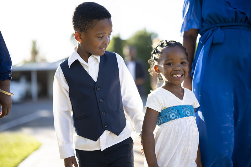 Two children smile at each other during a vow renewal ceremony. They're outdoors on a sunny day, wearing semi formal wedding clothes, standing next to a woman in a blue dress