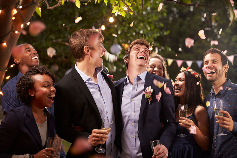 Two grooms laugh happily outdoors after their wedding ceremony while friends and family members raise a toast. It's a festive scene, with strings of lights twisted around the tree trunks and everyone smiling.