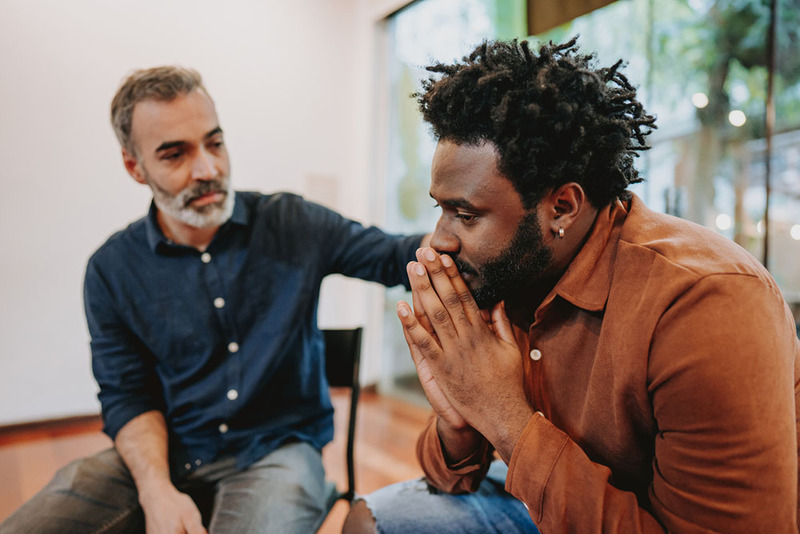 A funeral officiant rests a hand on a grieving man's shoulder during a meeting